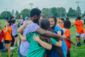 A group of young people wearing colorful shirts share a group hug outdoors on a grassy field. Other participants in bright orange, purple, red, and blue shirts are visible in the background, laughing, and interacting.