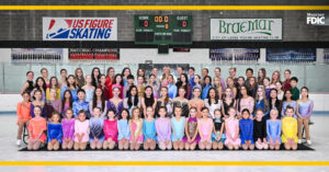 A group portrait of approximately 60 youth figure skaters in colorful, sparkling costumes posing on an indoor ice rink. They are arranged in tiered rows in front of a grey arena wall featuring a scoreboard and Braemar City of Lakes Figure Skating Club banners.